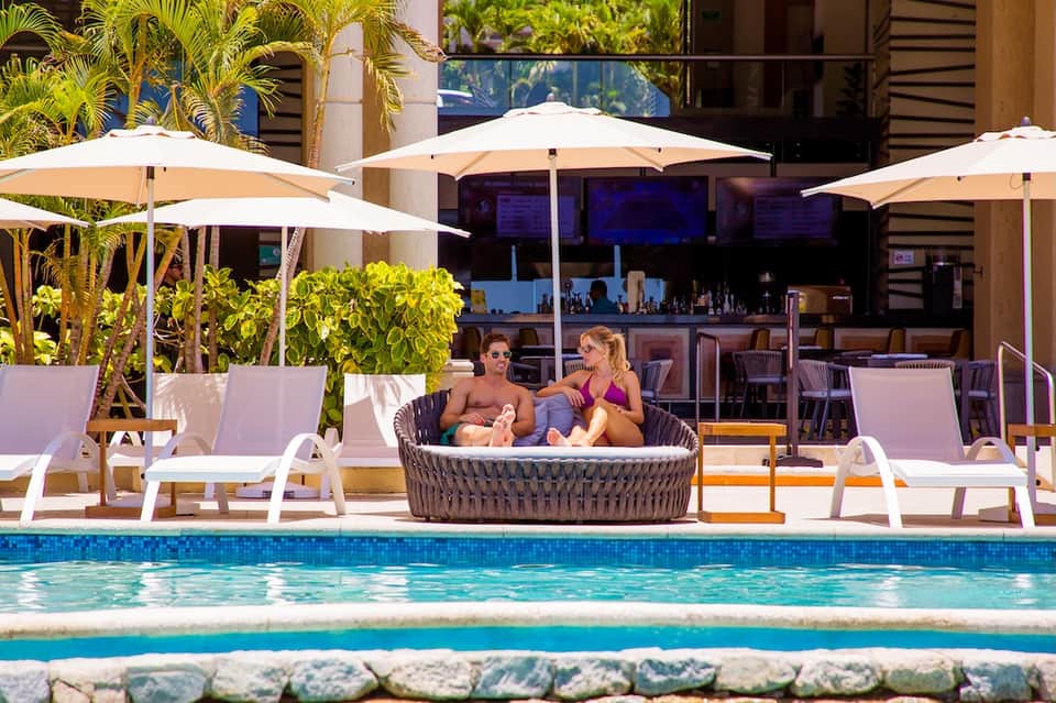 Pool deck with rattan lounge pod, white umbrellas, lounge chairs, and modern hotel building with ocean views
