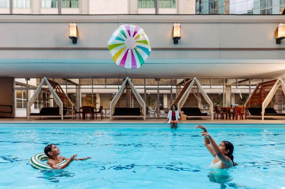 Indoor pool with swimmers, colorful umbrella, wooden cabanas, and large windows