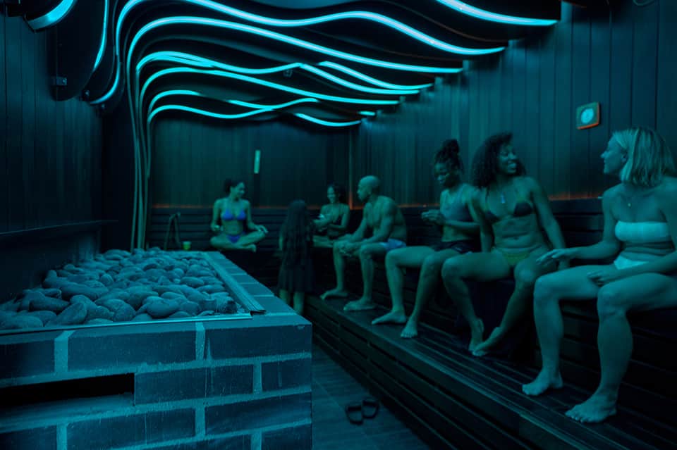 Spa sauna room with neon turquoise lighting and people seated on wooden benches