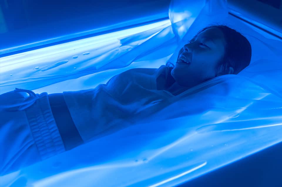 Person lying in blue-lit tanning bed with fluorescent UV light tubes glowing overhead