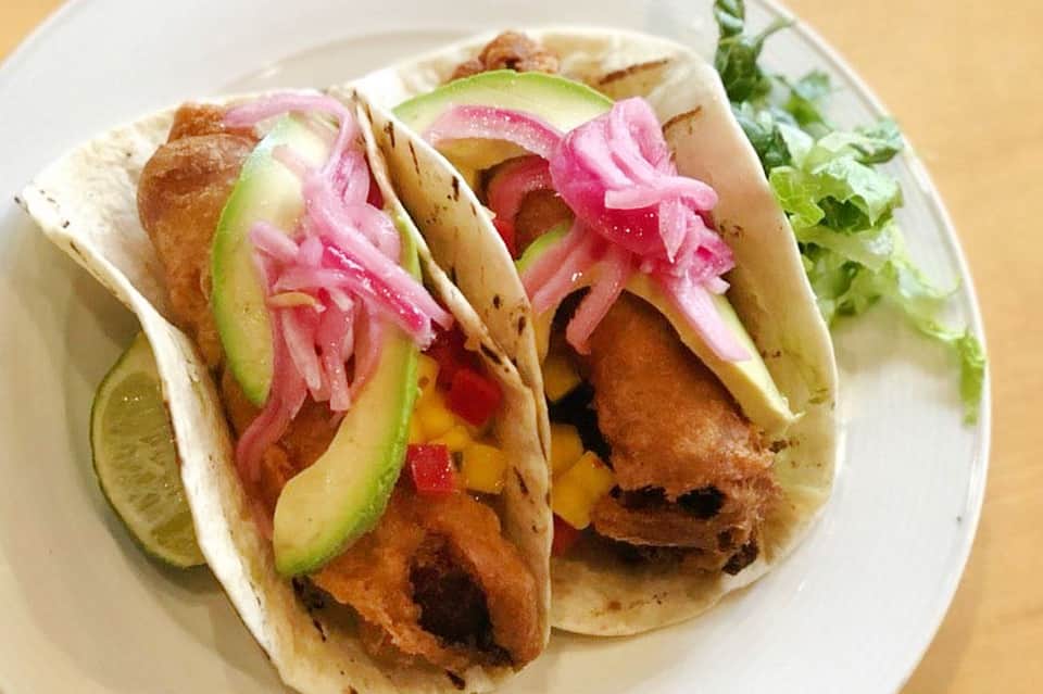 Two crispy fish tacos with pickled onions, avocado slices, and lime wedge on white plate