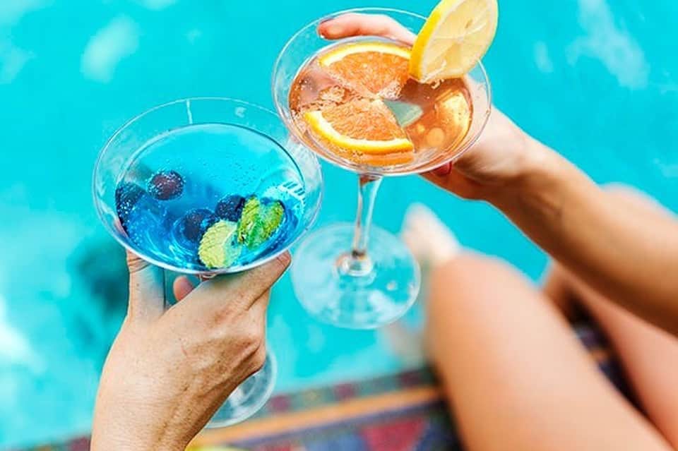 Two tropical cocktails with citrus garnish held up against bright turquoise pool water
