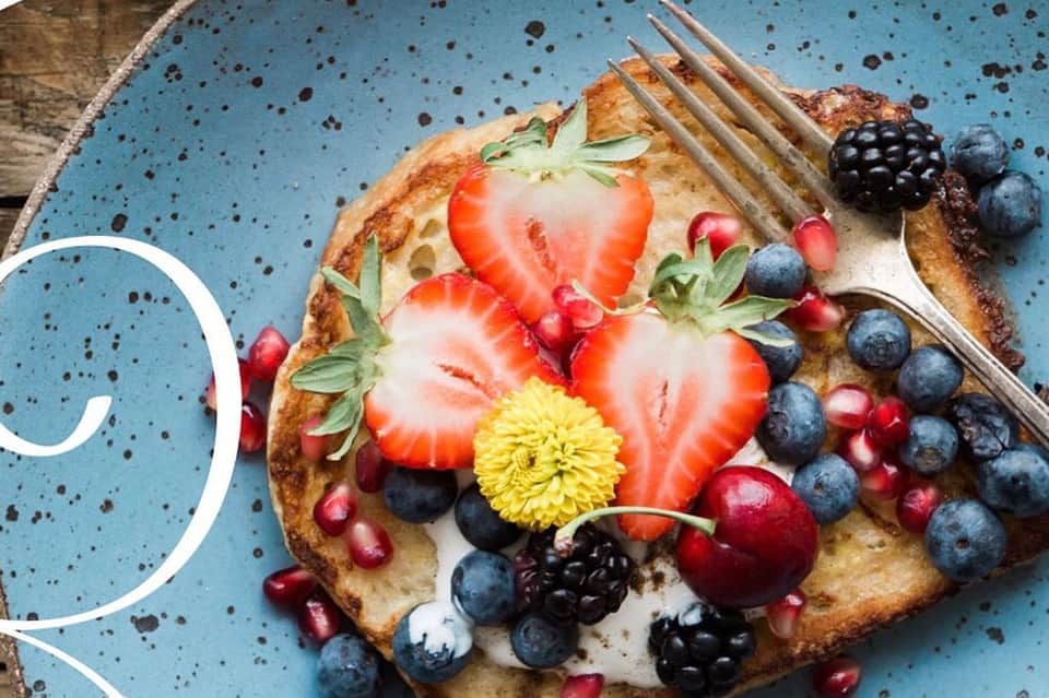 Gourmet toast topped with whipped cream, fresh strawberries, blueberries, blackberries, and yellow flower garnish on speckled blue plate