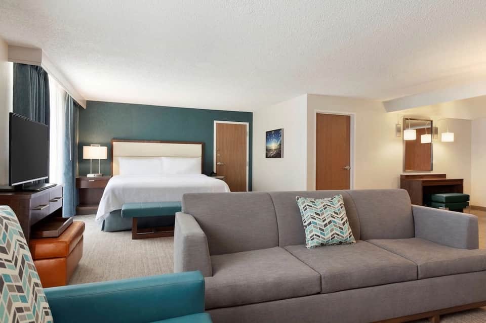 Hotel suite with teal accent wall, queen bed, sofa, and flat-screen TV in neutral tones