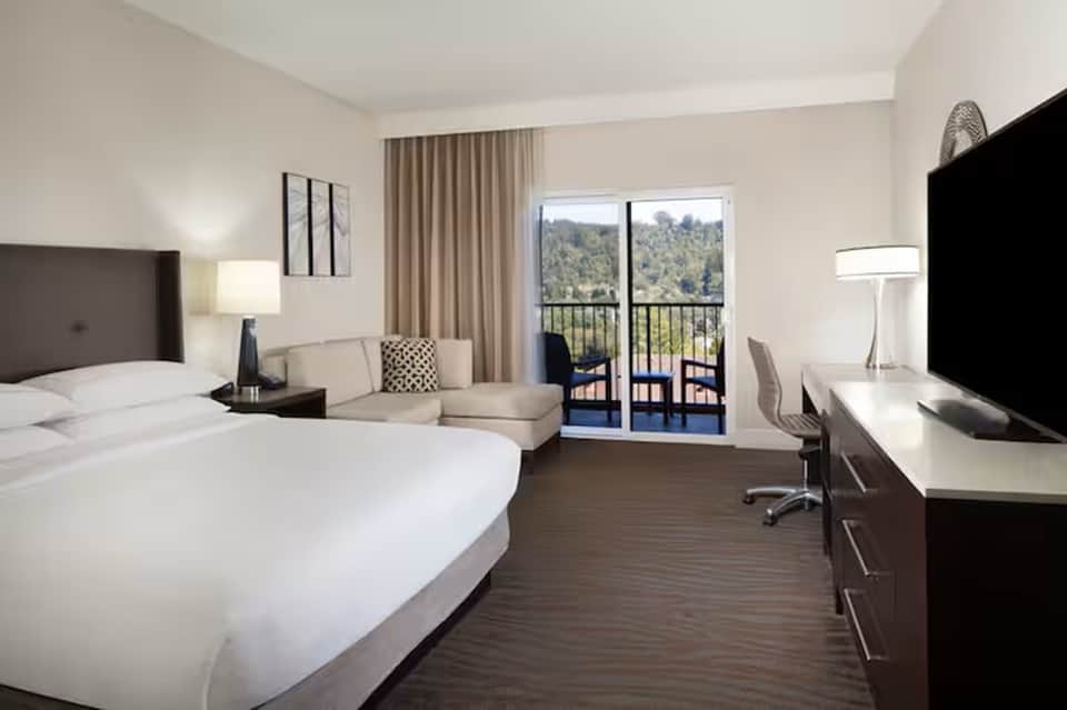 Contemporary hotel room with white bed, lounge seating, flat-screen TV, and balcony with mountain views