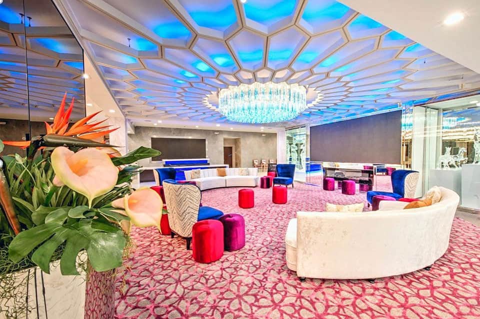 Luxurious hotel lobby with geometric ceiling, blue and pink accents, cream seating, and tropical flower arrangement