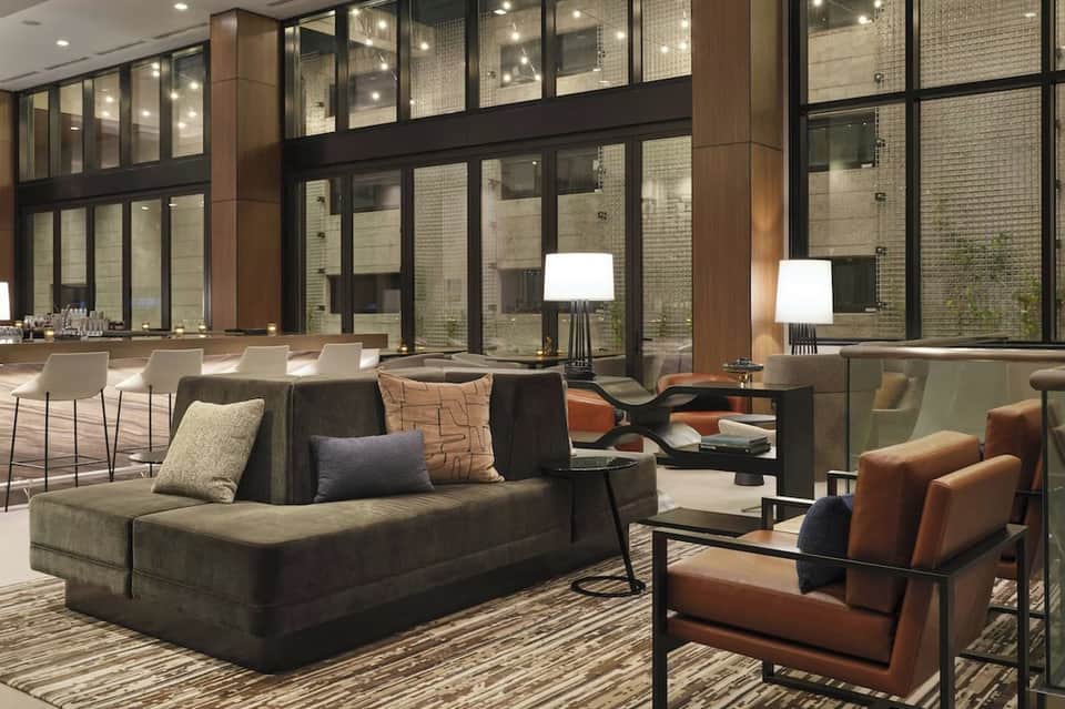 Contemporary hotel lounge with dark sofa, leather chairs, floor lamps, and floor-to-ceiling windows