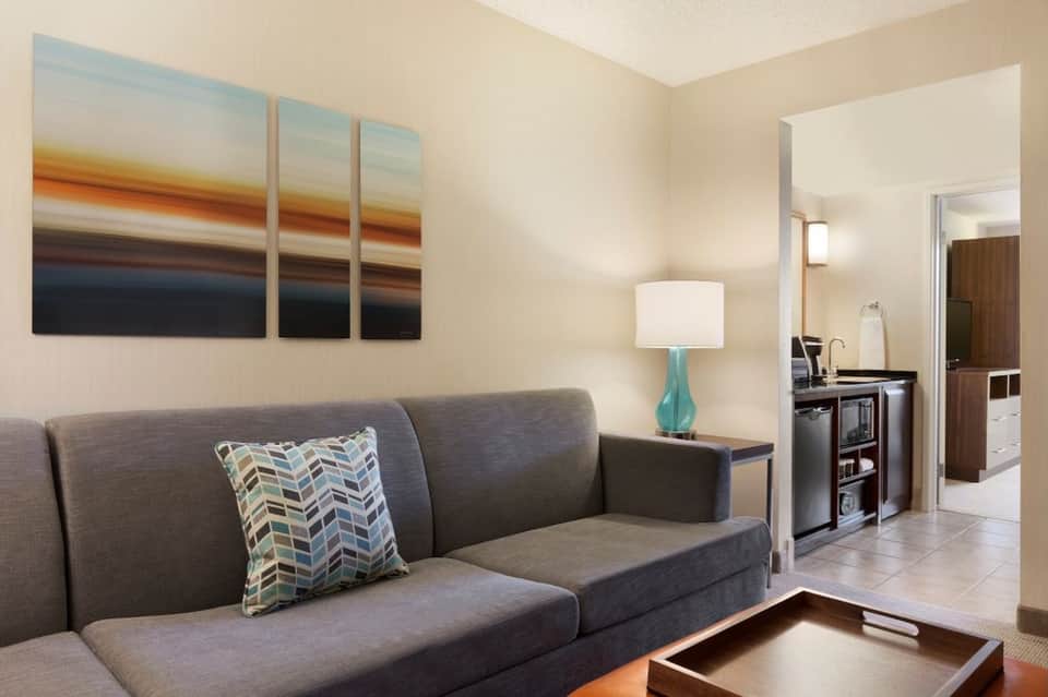 Hotel suite living area with gray sofa, turquoise lamp, and kitchenette