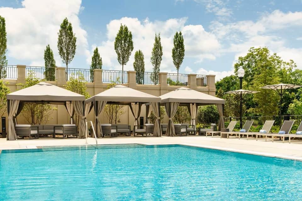 Resort pool with beige cabanas featuring curtains, tall cypress trees, and landscaped grounds