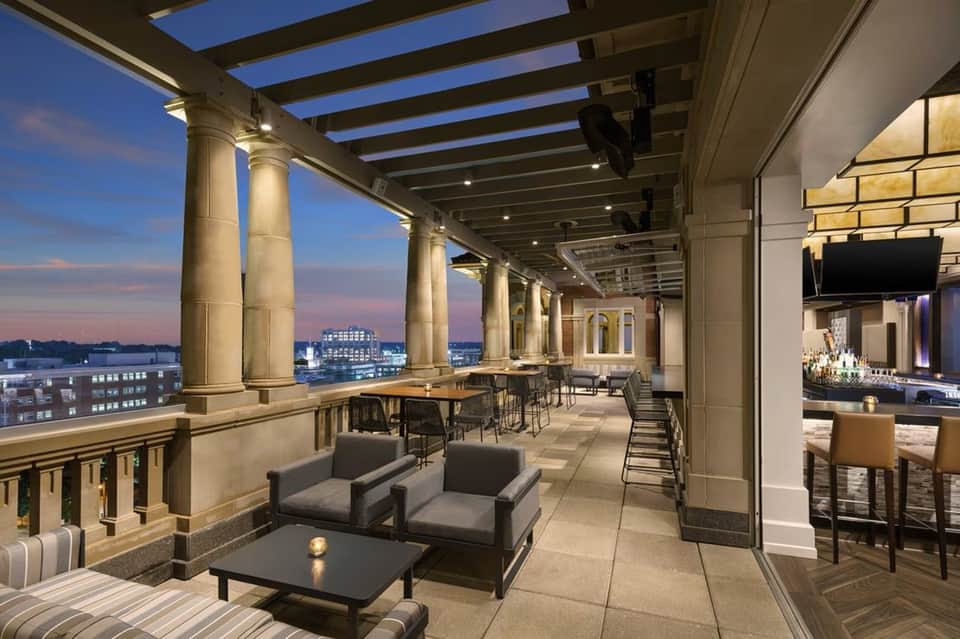 Upscale rooftop terrace at dusk with columns, lounge seating, bar area, and city skyline views