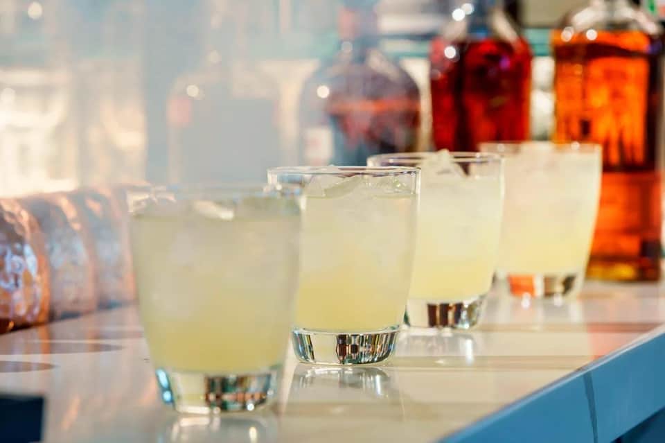 Row of clear glasses with pale yellow cocktails on a bar with blurred bottles in background