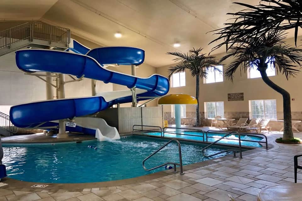 Indoor water park with blue spiral waterslide, pool, palm tree, and high ceiling with windows