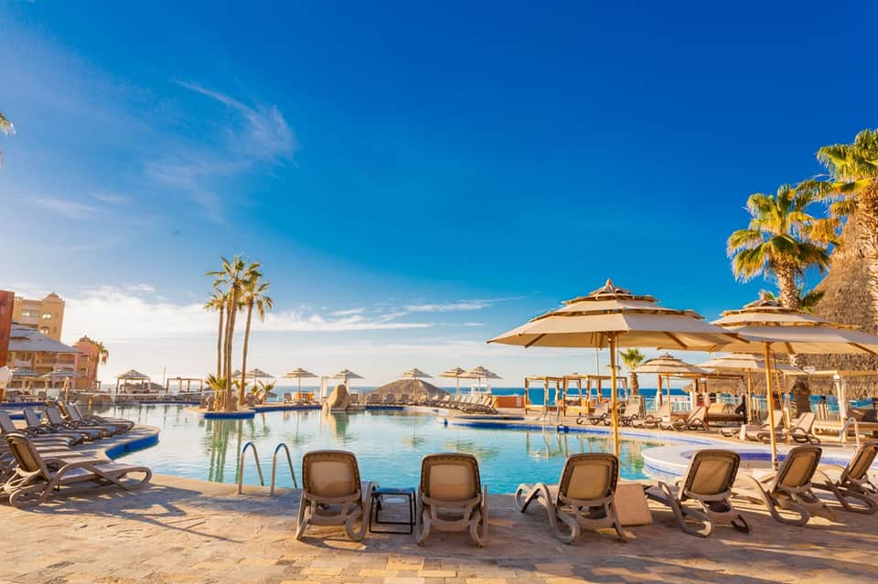 Tropical resort pool complex with lounge chairs, umbrellas, palm trees, and ocean views at day