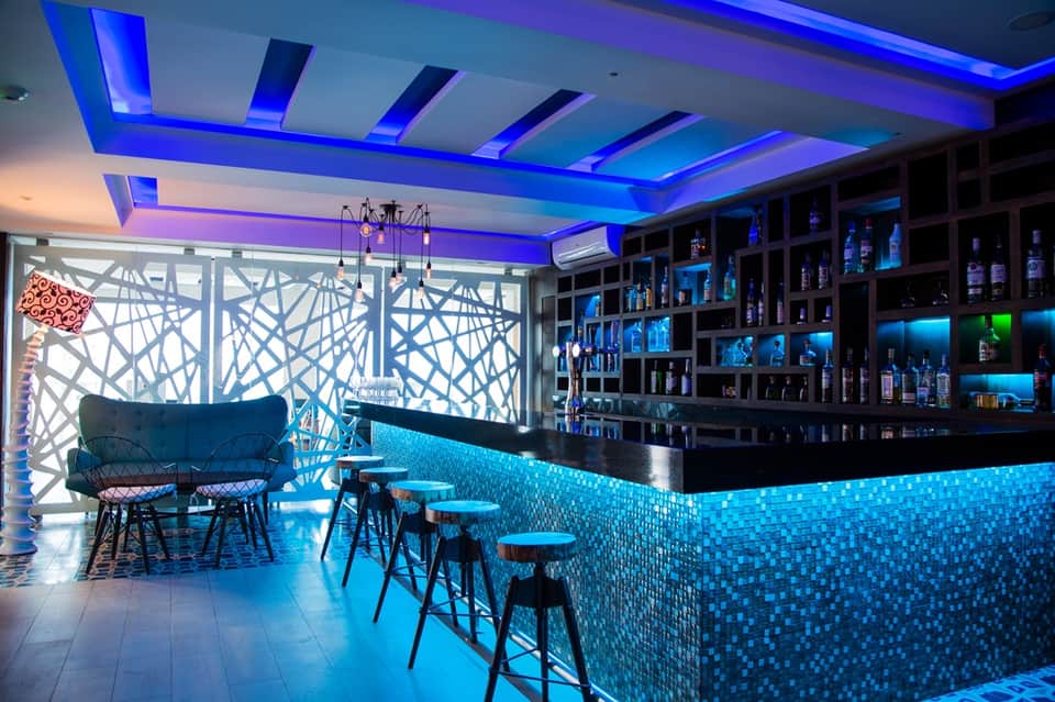 Modern hotel bar with blue LED ceiling, black bar counter, patterned backsplash, and illuminated shelving