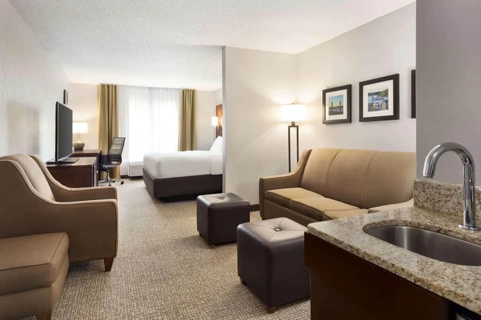 Spacious hotel suite featuring bed, sitting area with tan leather sofa, kitchenette with granite countertop, and flat-screen TV.
