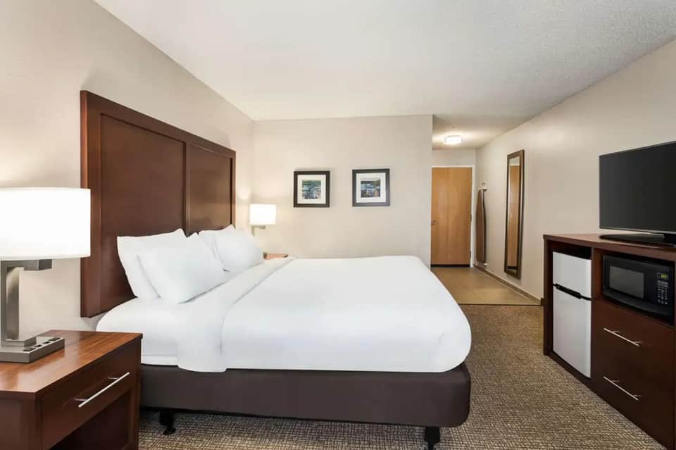 Hotel bedroom with queen bed, wood headboard, TV, desk, and warm neutral color palette