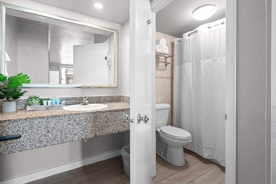 Clean hotel bathroom with granite countertop, large mirror, toilet, and shower with white curtain