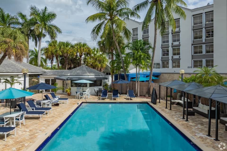 Resort pool with turquoise water, palm trees, lounge seating areas, cabanas, and multi-story hotel building in background