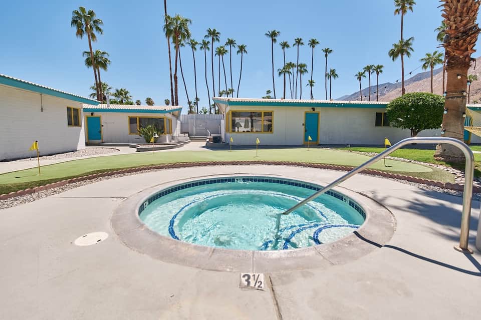 Mid-century turquoise and white resort buildings with hot tub, palm trees, and desert mountain backdrop