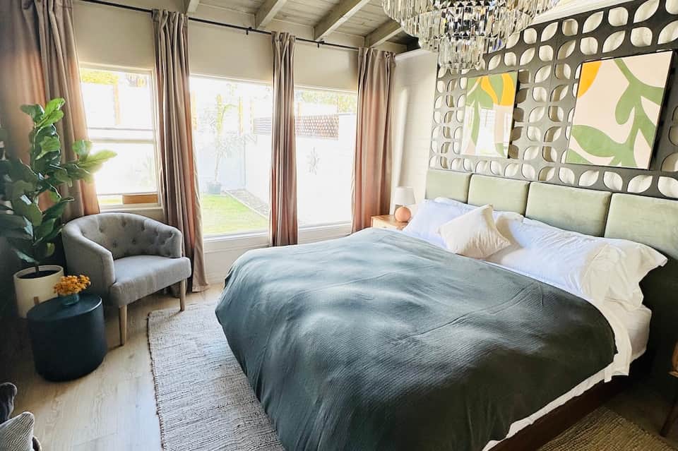 Bedroom with large windows, gray bedding, decorative mirror, and chair overlooking garden