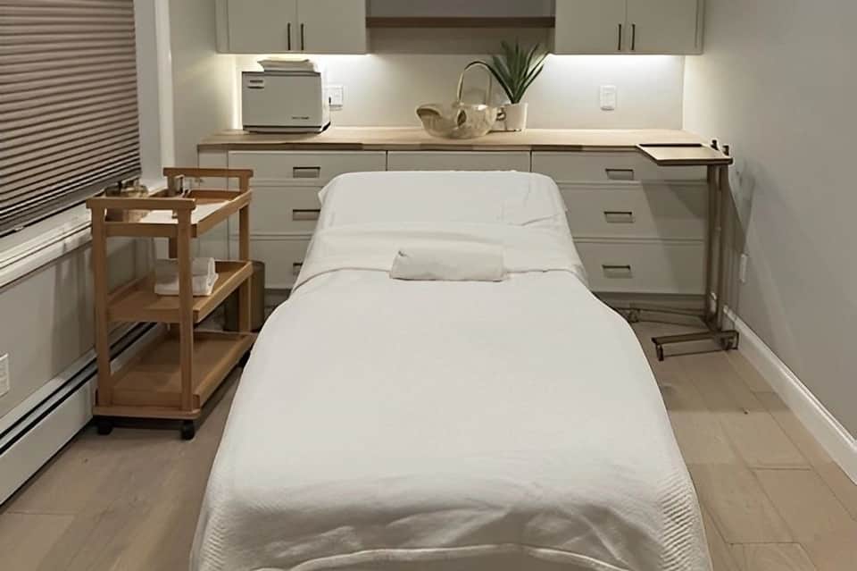 Minimalist spa treatment room with massage bed, vanity, wooden shelving, and clean white decor