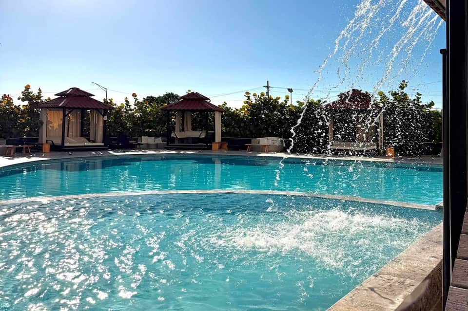 Turquoise pool with water feature fountain, dark umbrellas, and ivy-covered walls under clear sky