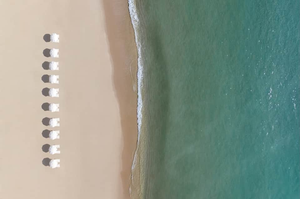 Aerial view of pristine sandy beach with rows of white umbrellas creating geometric shadows in turquoise shallow water