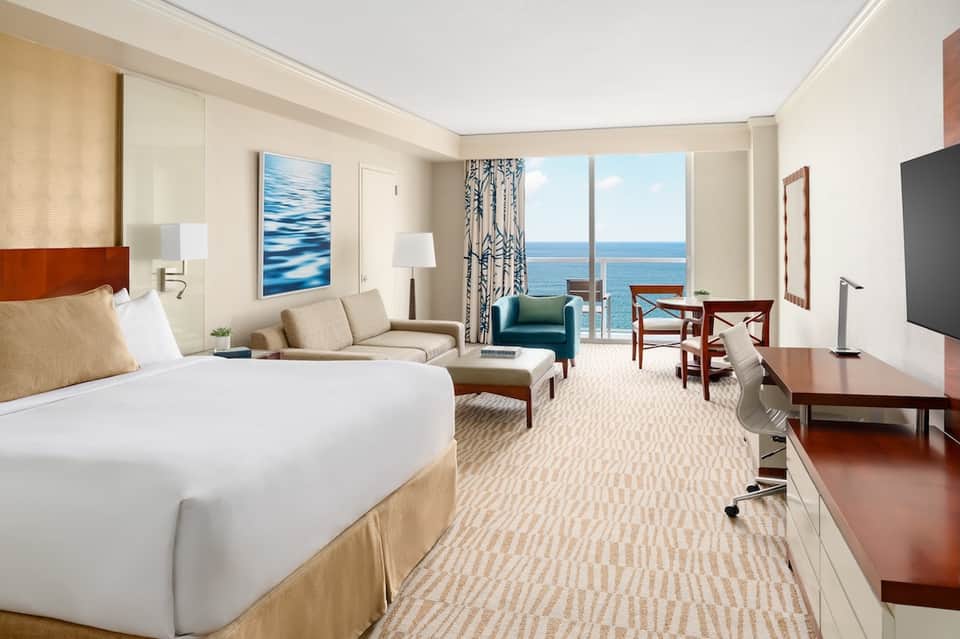 Luxury oceanfront hotel room with king bed, sofa seating, floor-to-ceiling windows, and sea views