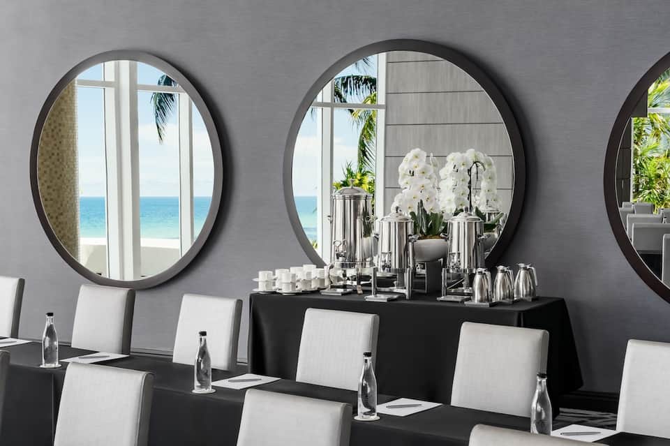 Beachfront meeting room with large round mirrors, black conference table, white chairs, and ocean views
