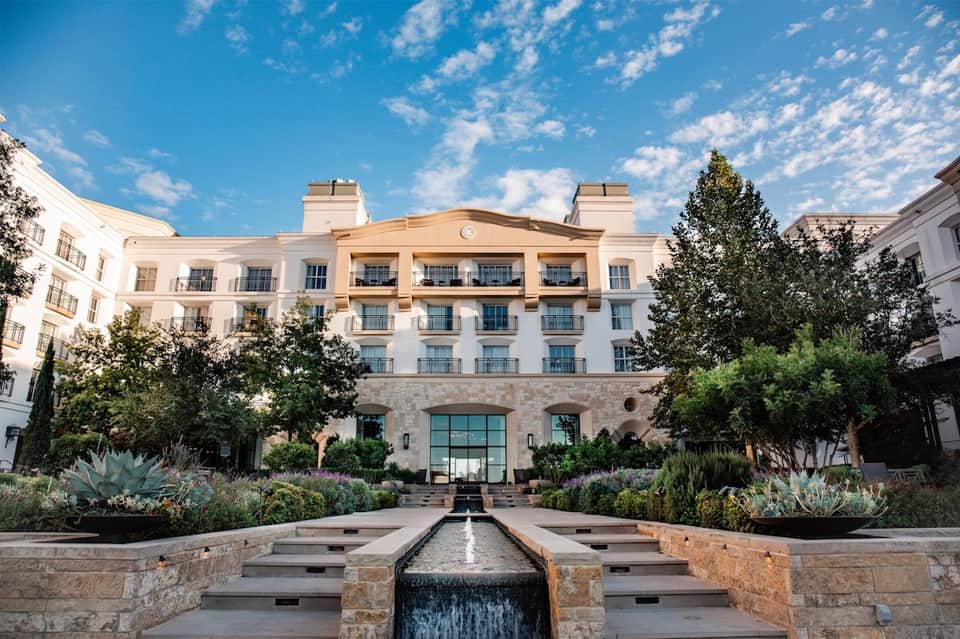 Elegant hotel courtyard with white and tan facade, manicured gardens, succulent plantings, and water feature.