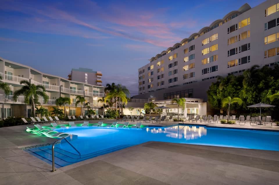 Illuminated resort pool at dusk with lounge chairs and multi-story hotel buildings