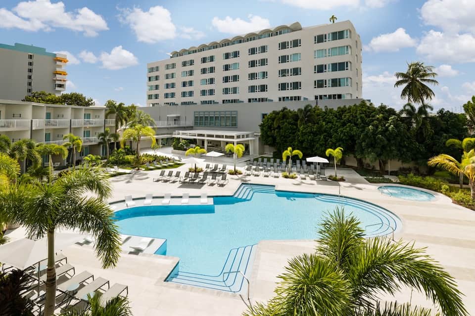Beachfront resort with multi-story hotel building, curved swimming pool, palm trees, and lounge chairs