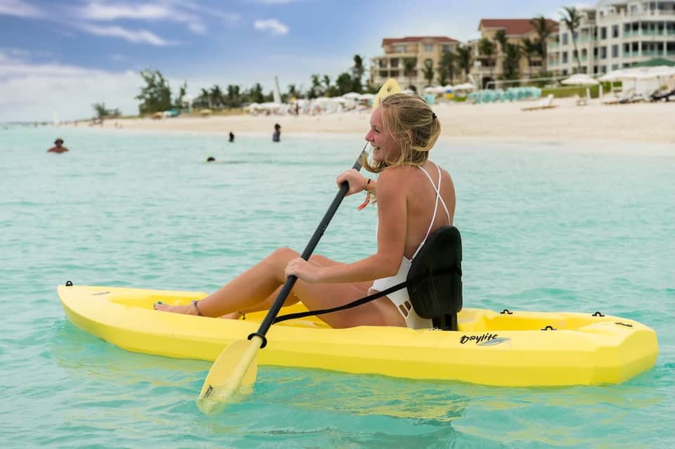 Woman kayaking in clear turquoise ocean water with beachfront resort buildings visible in background