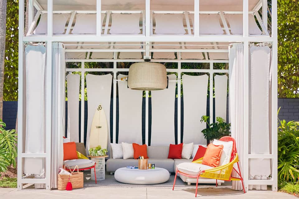 Modern pergola lounge with gray sofa, red cushions, hanging pendant light, and surfboard decor