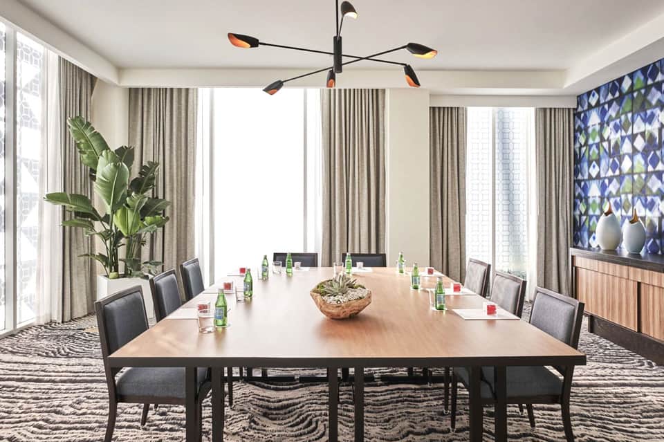 Modern conference room with long wooden table, gray upholstered chairs, floor-to-ceiling windows, and contemporary chandelier