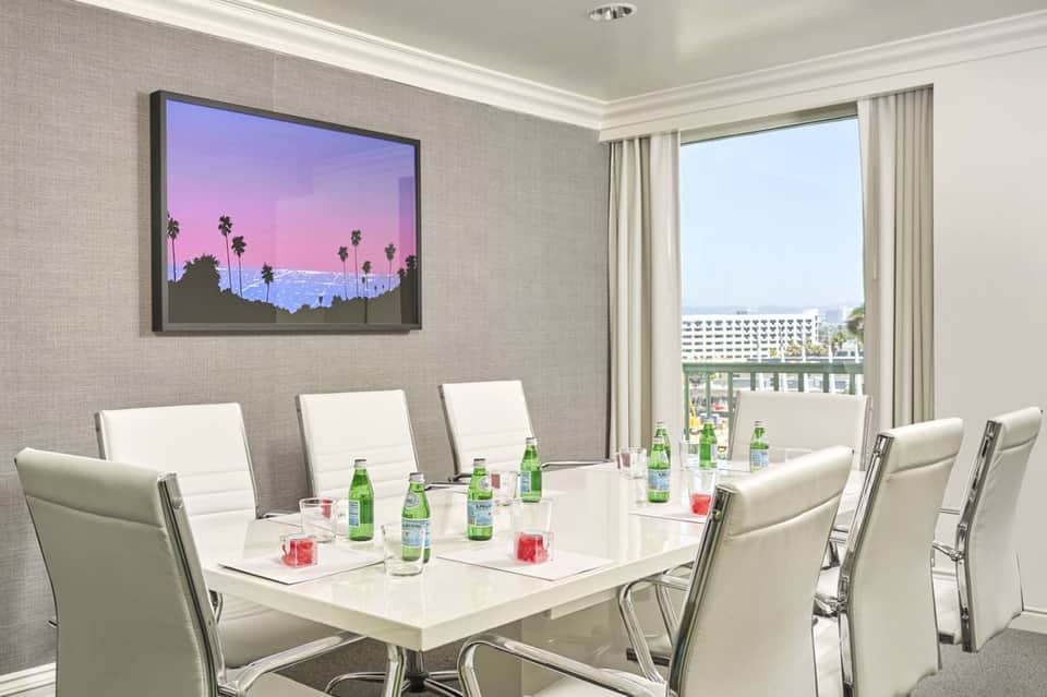 Hotel conference room with white table, white leather chairs, palm tree artwork, and city view from floor-to-ceiling windows