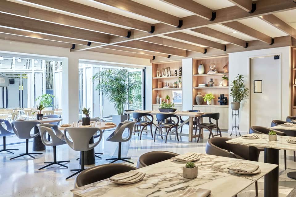 Modern restaurant dining area with marble tables, curved chairs, plants, and glass doors