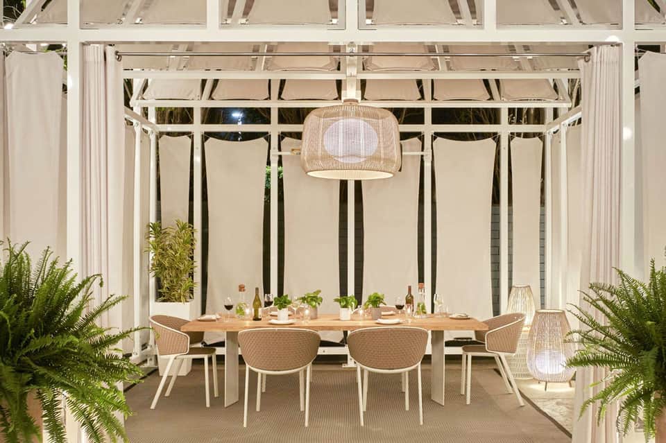 Modern outdoor dining pavilion with wooden table, woven chairs, and neutral shade curtains