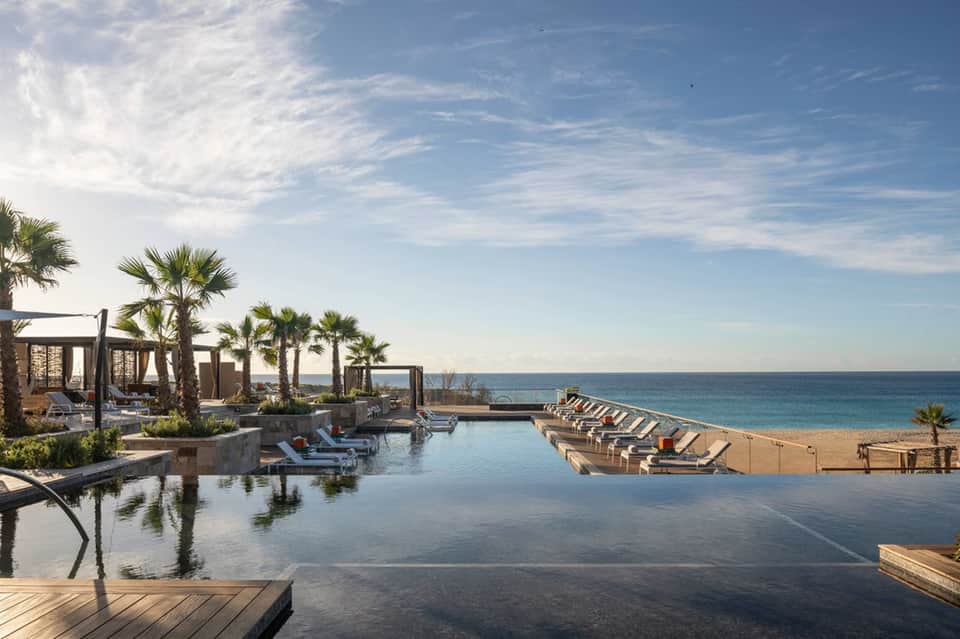 Beachfront infinity pool overlooking ocean, palm trees, cabanas, and sandy beach with loungers