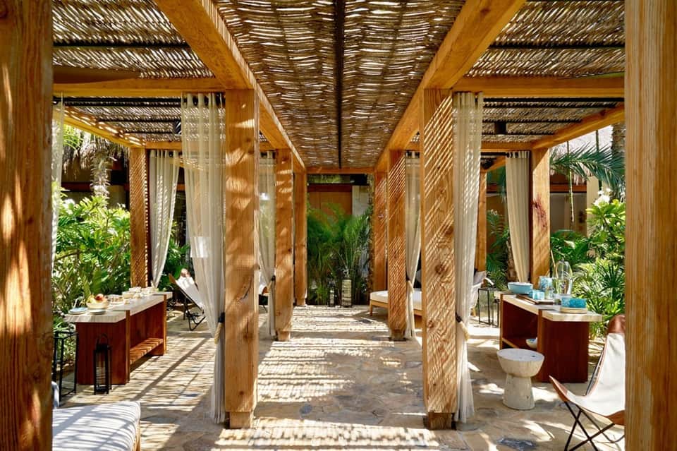Covered spa pavilion with wooden pergolas, flowing curtains, red vessels, and tropical plants