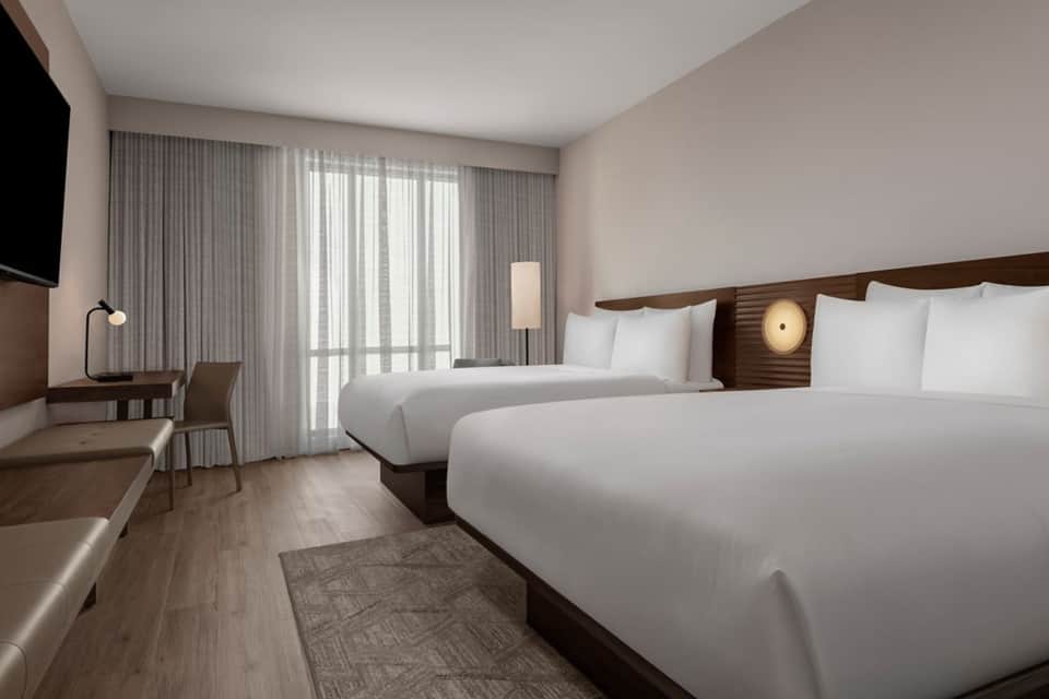 Modern hotel room with dual beds, wood headboard, floor-to-ceiling curtains, and desk area