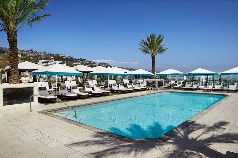 Resort rooftop pool with blue water, turquoise umbrellas, white loungers, and mountain views