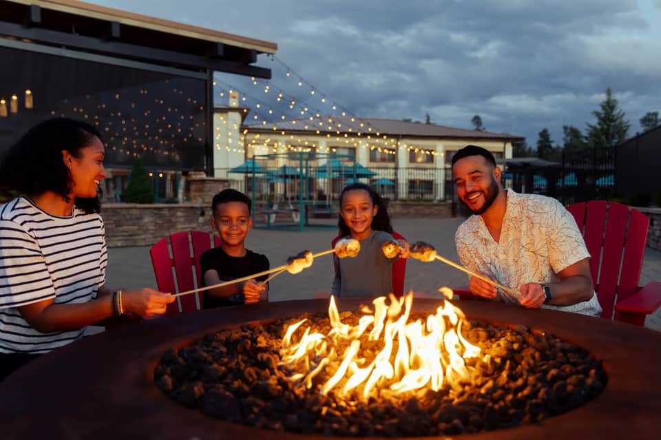 Family roasting marshmallows around fire pit at dusk with resort building and string lights in background