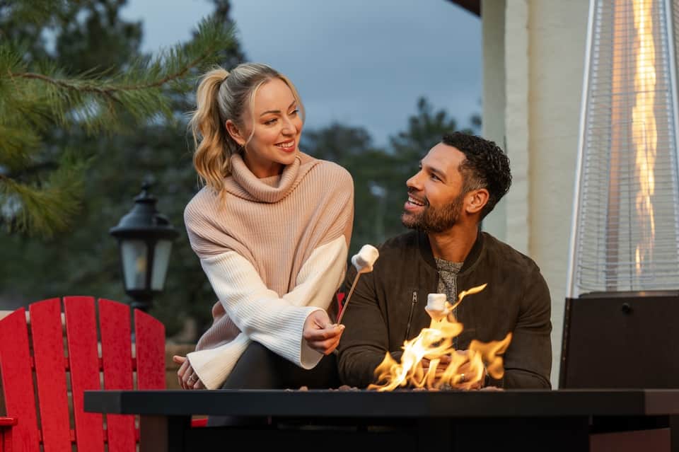 Couple by fire pit at night with s'mores, pine trees, and warm ambient lighting