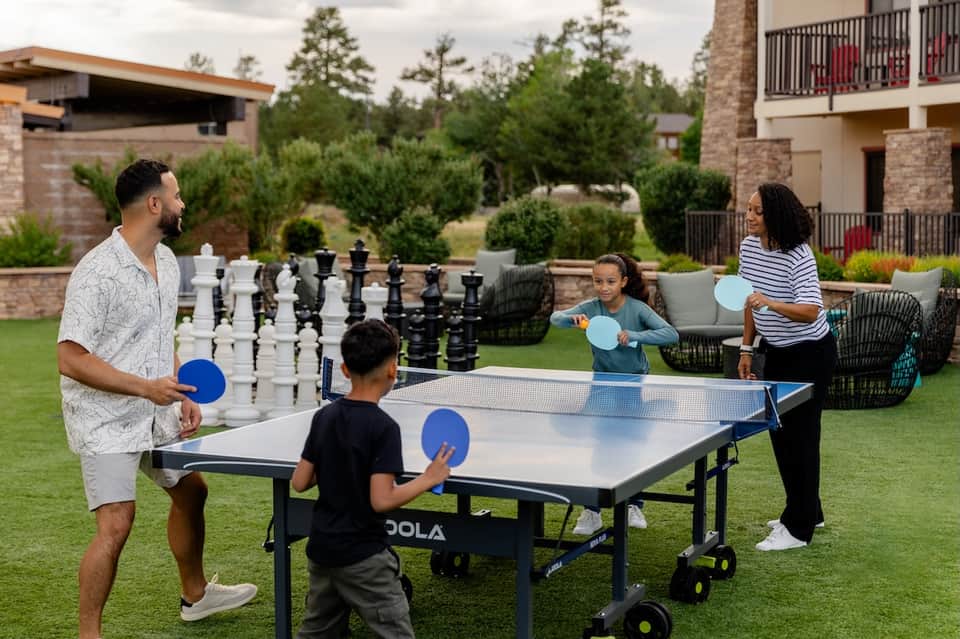 Family playing table tennis and chess on resort lawn with brick buildings and manicured landscaping