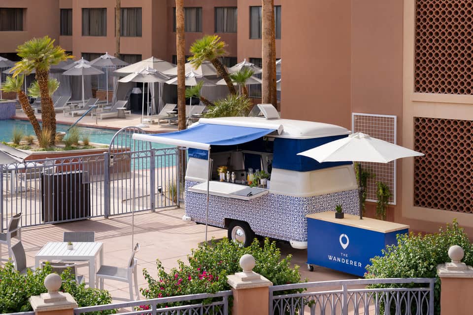Resort poolside with blue and white food cart, white umbrellas, palm trees, and terracotta building