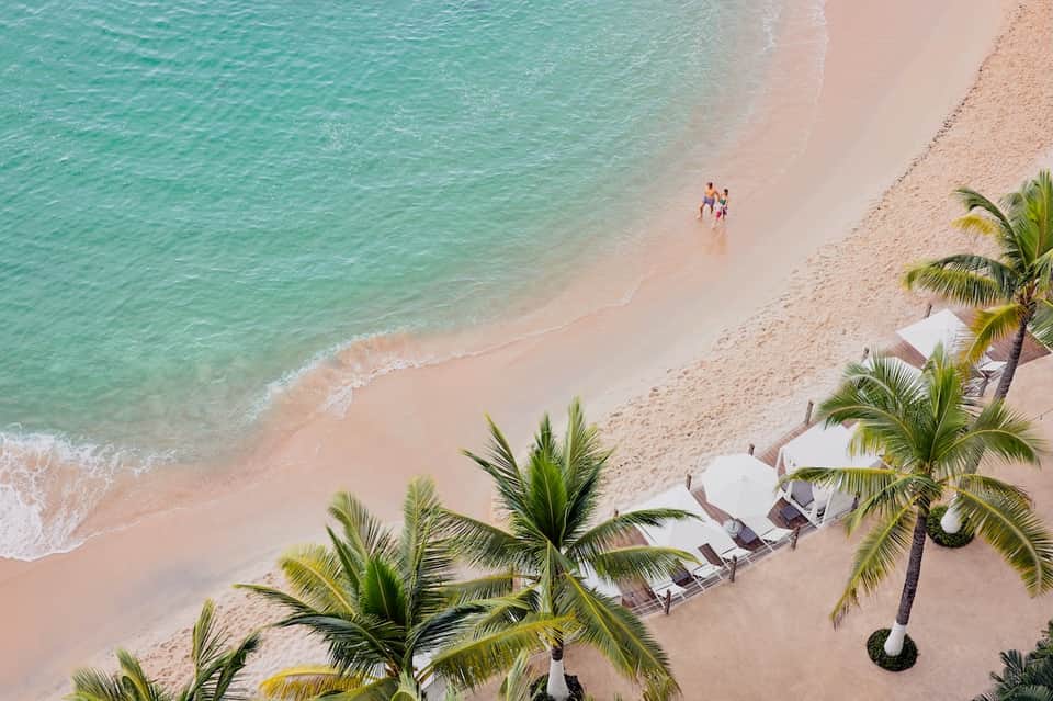 Aerial beach view with turquoise waters, pristine sand, palm trees, and beachfront lounge chairs in a tropical setting