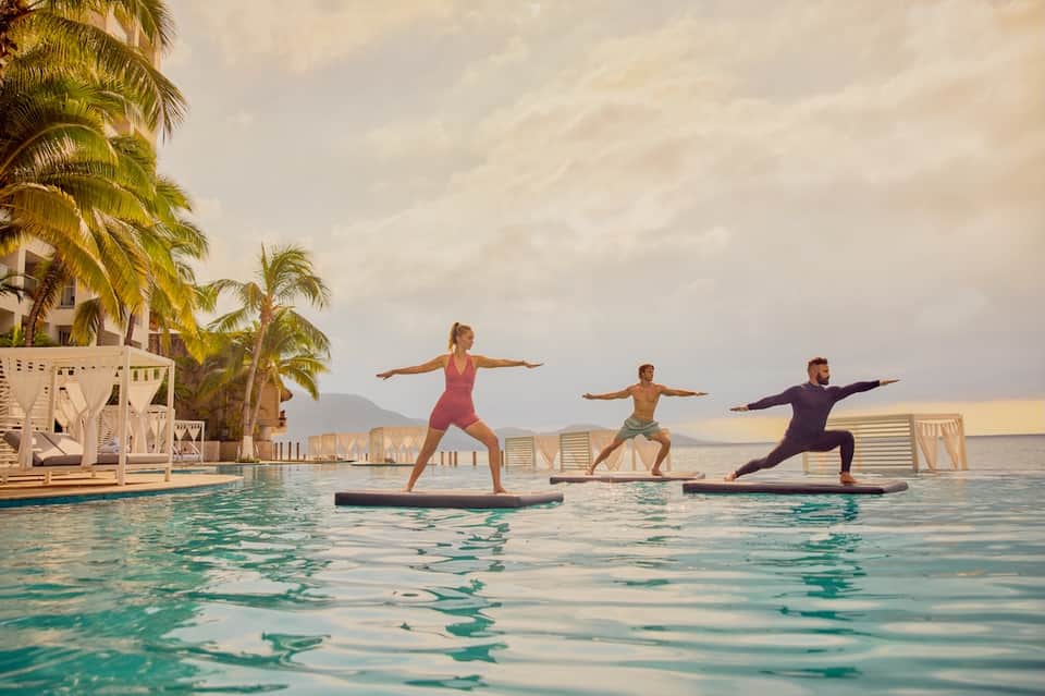Outdoor water yoga class with three instructors and participants on floating platforms in tropical bay