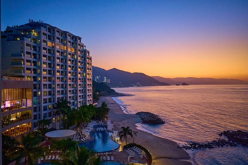Beachfront resort at dusk with illuminated pool, palm trees, and mountains across the bay
