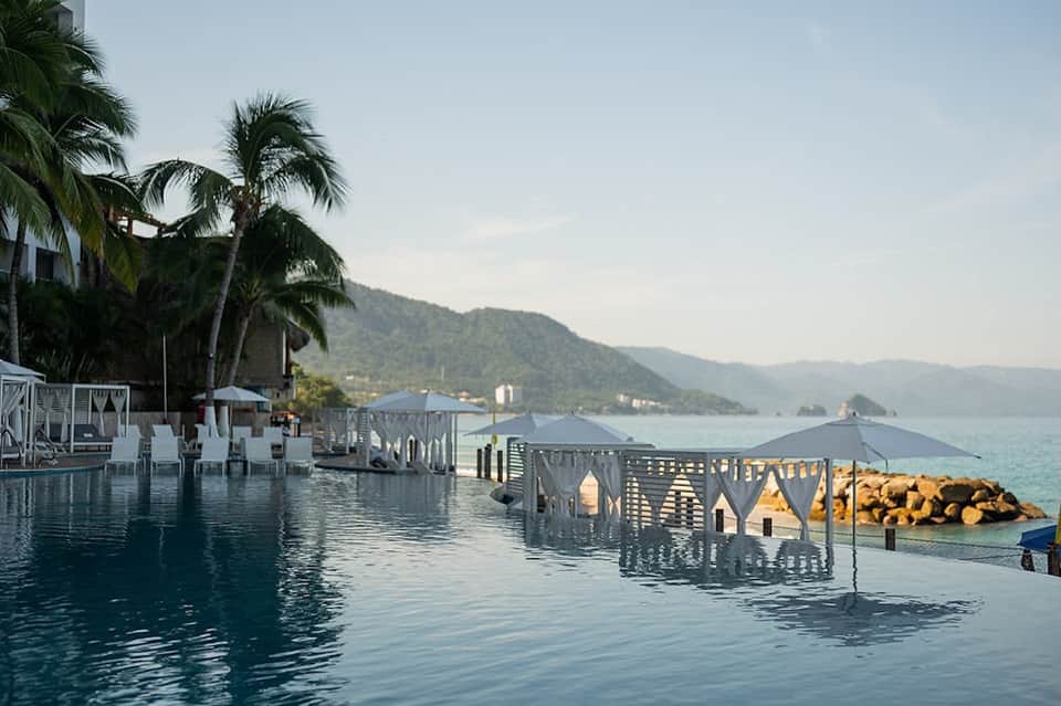 Infinity pool merging with ocean, palm trees, mountain views, and white cabanas at sunset
